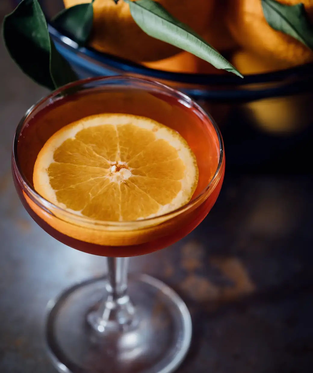 A rosy cocktail in a coupe with an orange wheel, a bowl of oranges behind it on the dark bar