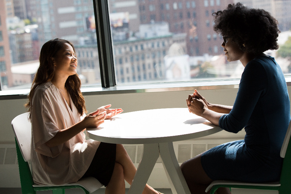 Two women talking across a small round table beside a tall office window