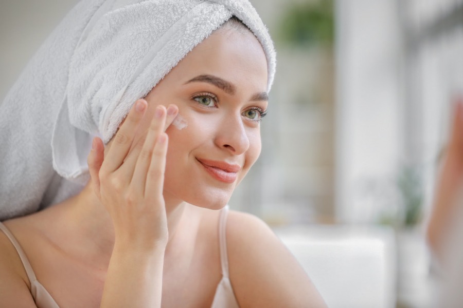 Woman with a towel on her hair smoothing cream onto her cheek at home