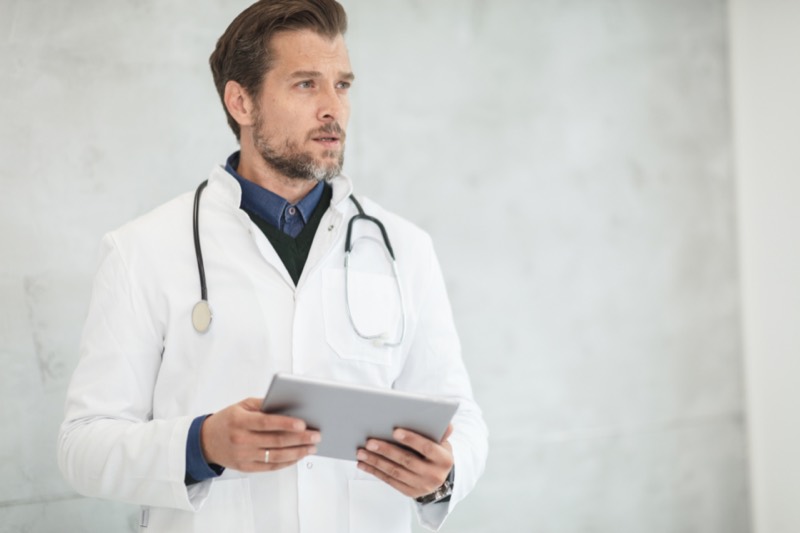 Marcus Lund in a white coat with a stethoscope, holding a tablet.