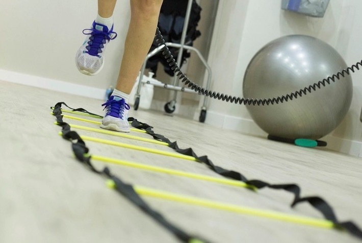 A patient steps through an agility ladder in the rehab gym, with an exercise ball nearby.