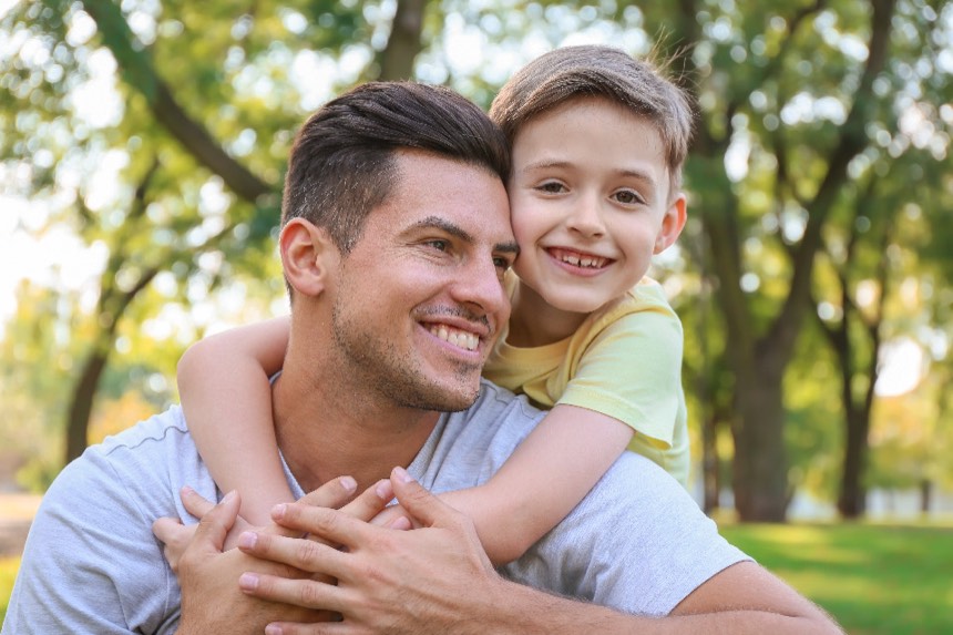 A father and his young son hug in a park; the boy grins with a missing front tooth