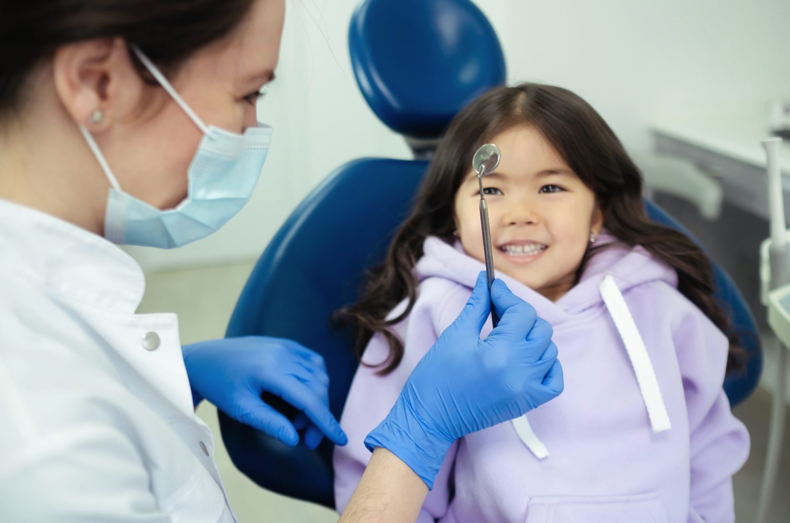 A smiling girl in a blue dental chair watches the dentist hold up a small mirror