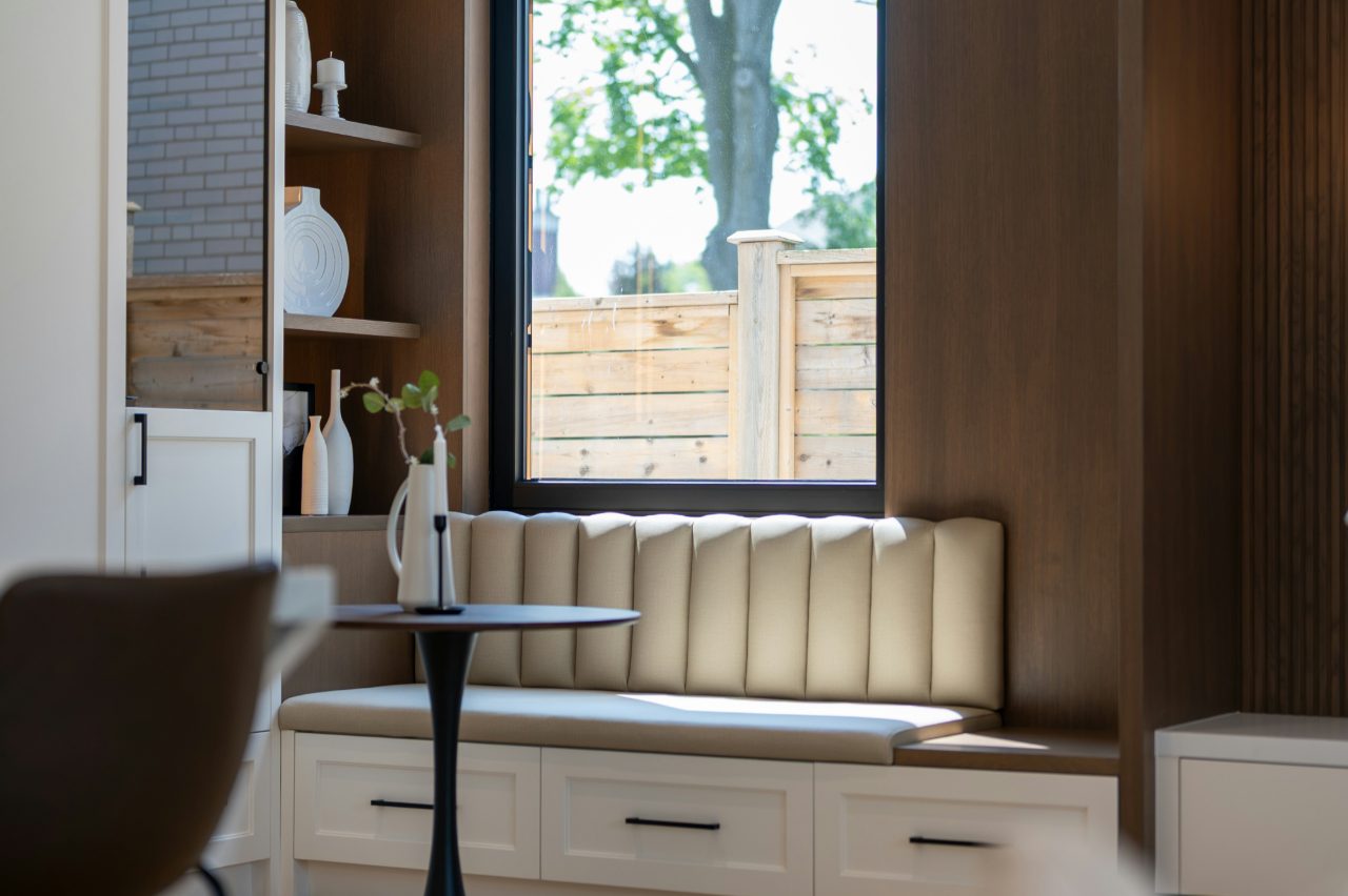 Kitchen nook with a channel-tufted window seat over built-in drawers, open shelving and a small round table beside a tall window