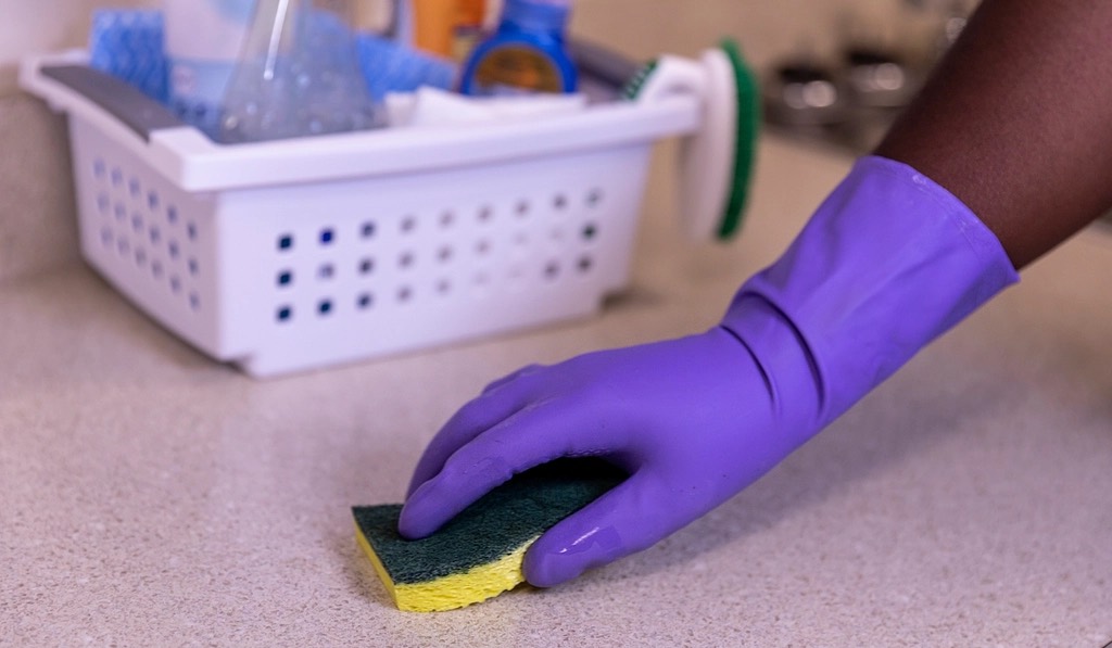 A gloved hand wiping a kitchen counter with a sponge, a caddy of supplies behind