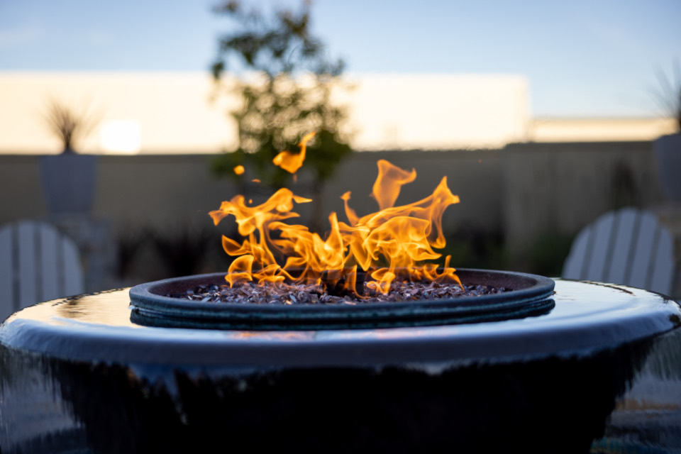 Flames rising from a dark stone fire bowl at dusk, with Adirondack chairs blurred behind it