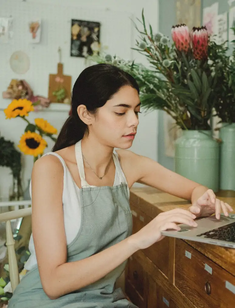 A florist checking orders on a laptop among sunflowers and proteas