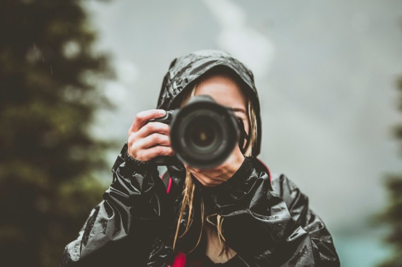 Maren in a black rain jacket with the hood up, raising her camera among fir trees