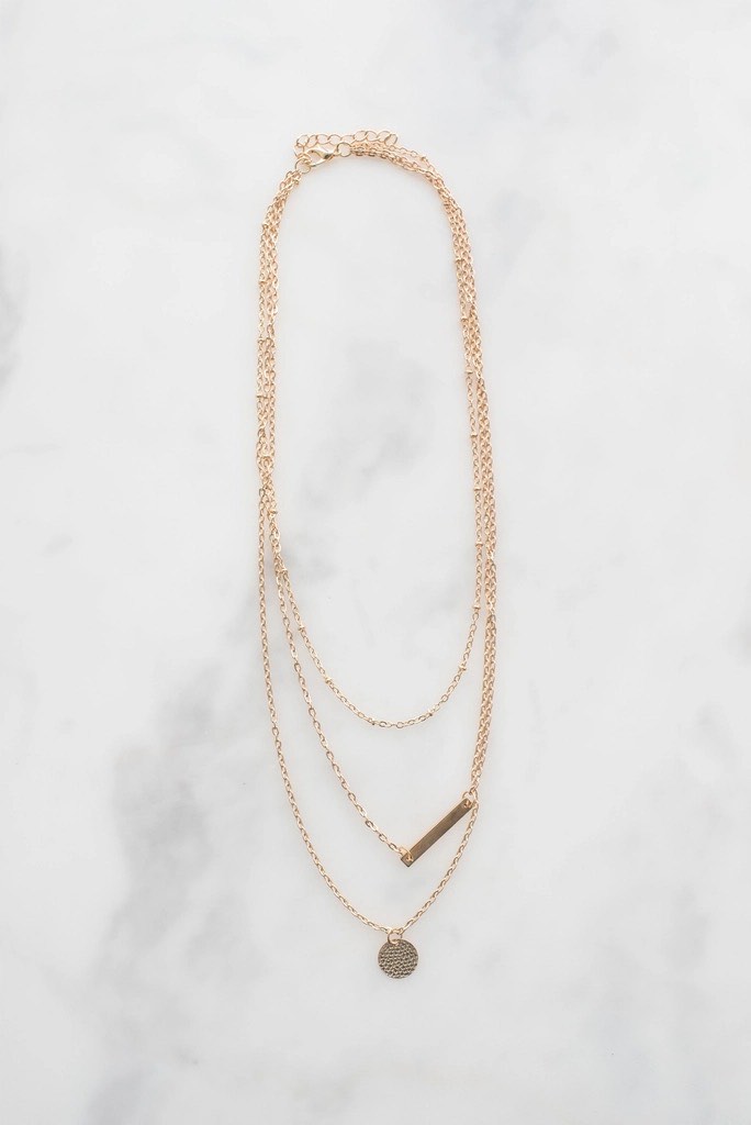 Three fine gold chains laid out on white marble, one with a small bar and one with a hammered disc