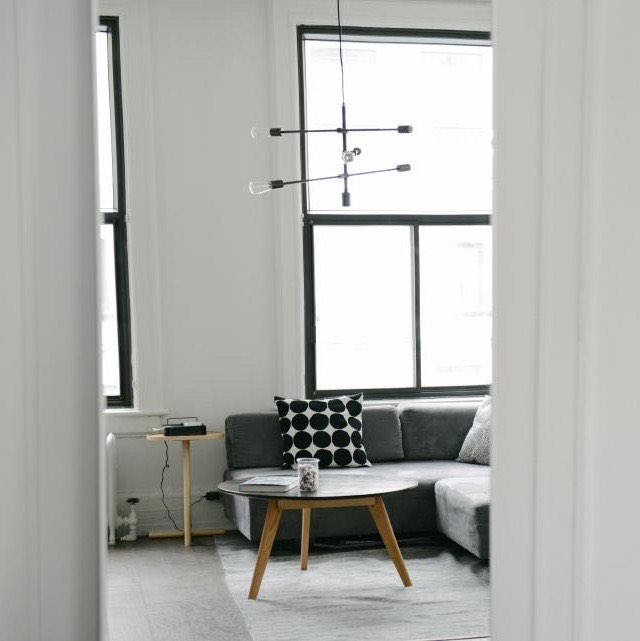 White living room with tall black-framed windows, a grey sectional sofa, a round wooden coffee table and a slim brass pendant