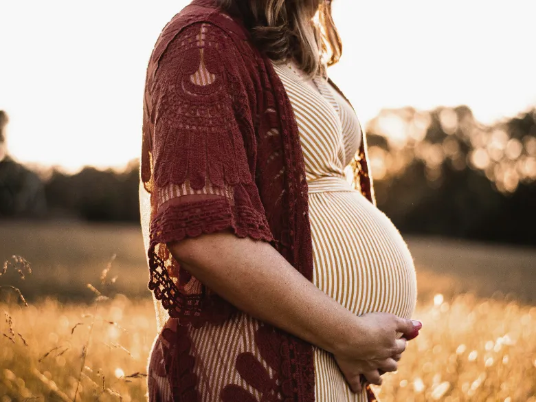 A pregnant woman in a striped dress and red cardigan cradling her belly in a golden field