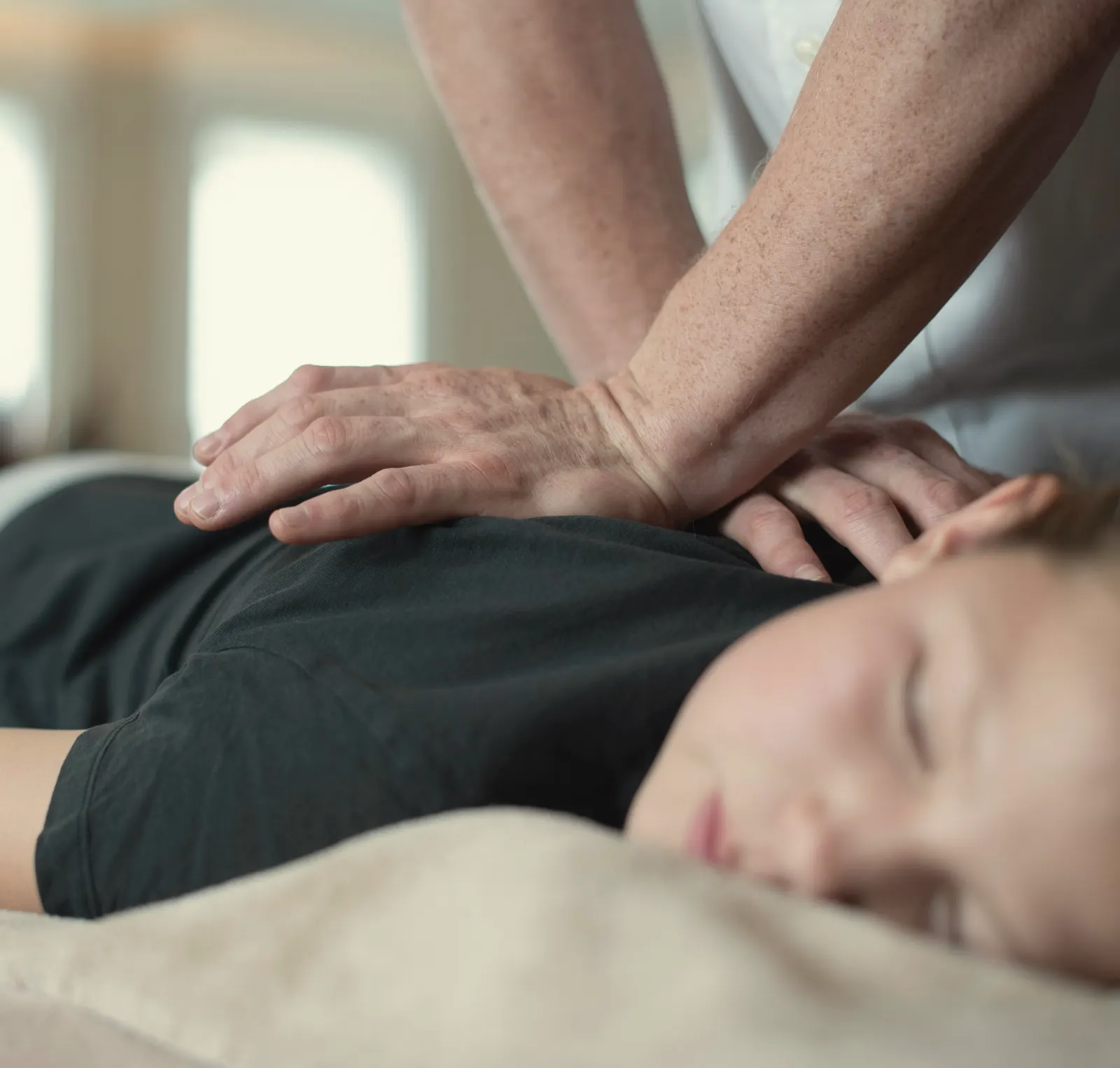 A practitioner's hands resting on a client's upper back while she lies face down, eyes closed, in soft window light