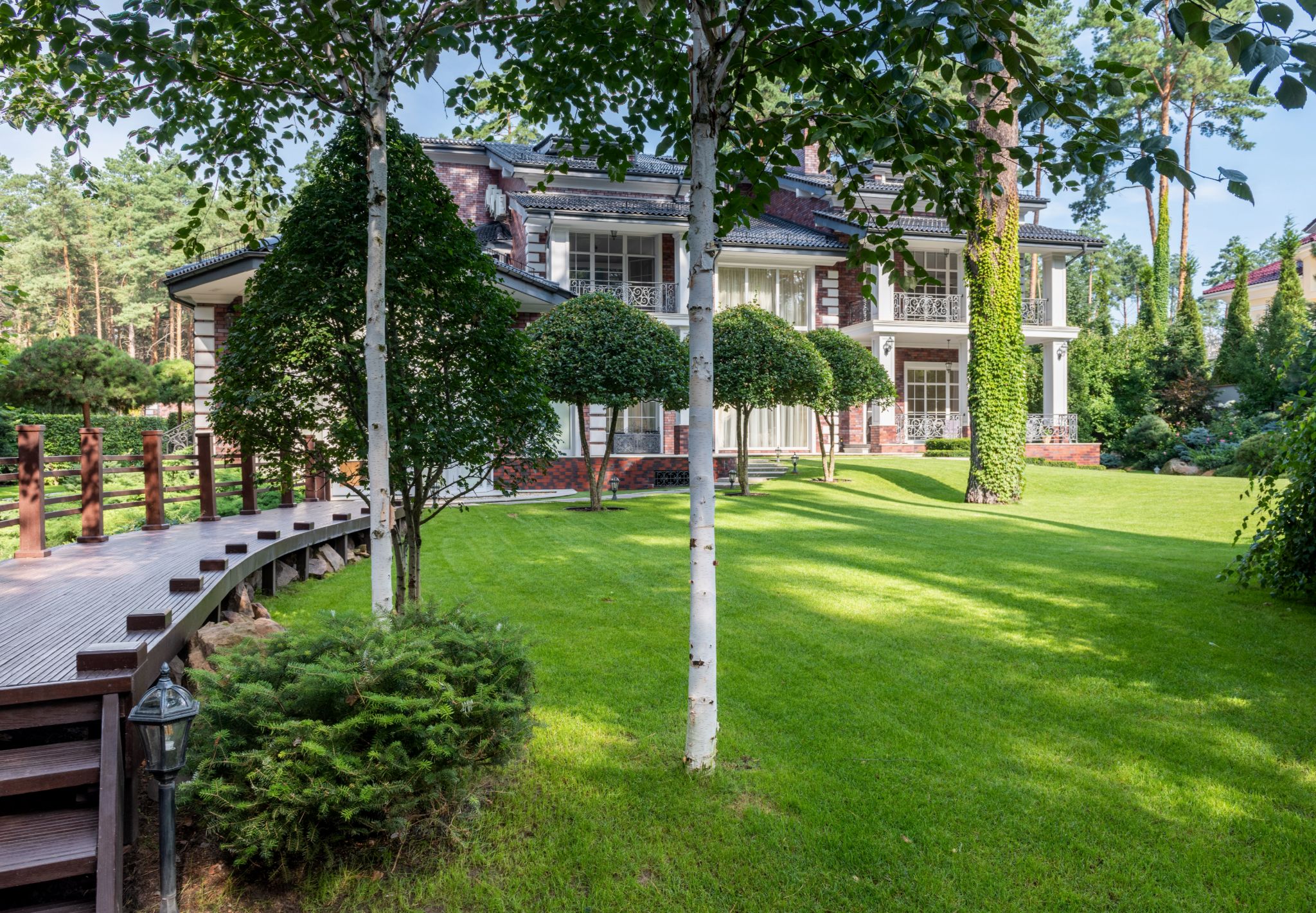 A brick house behind young white birches, with a clipped lawn, round-headed standard trees and a curving timber boardwalk