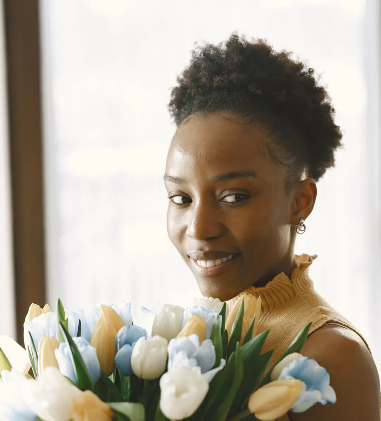 Imani Okafor, the owner, smiling over an armful of white, cream and pale blue tulips by a sunny window.