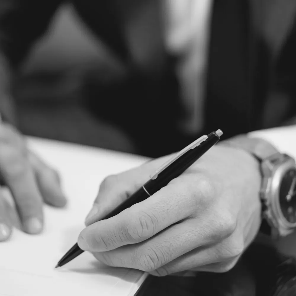 A hand in a shirt cuff signing a page with a black pen