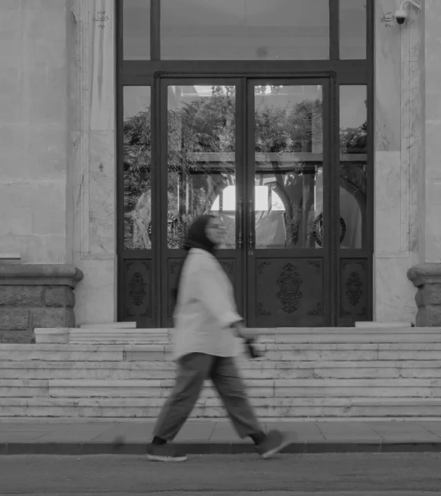 A woman walking past the stone steps and tall glass doors of an old building