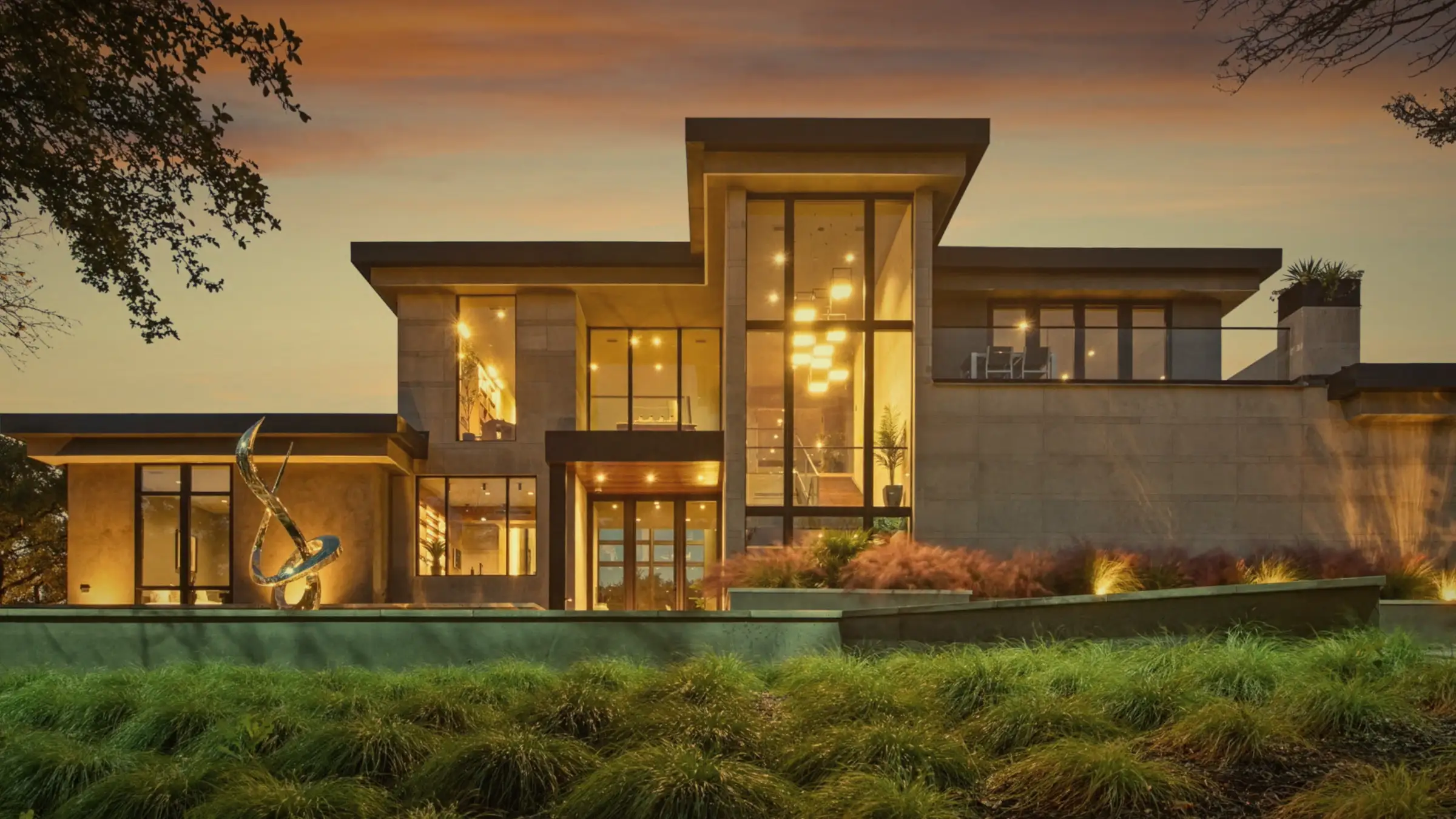 A modern stone and glass home at dusk, its tall windows glowing warm under an orange sky.