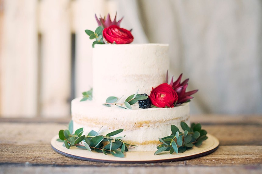 A two-tier naked cake with white frosting, decorated with red ranunculus, protea and eucalyptus leaves.