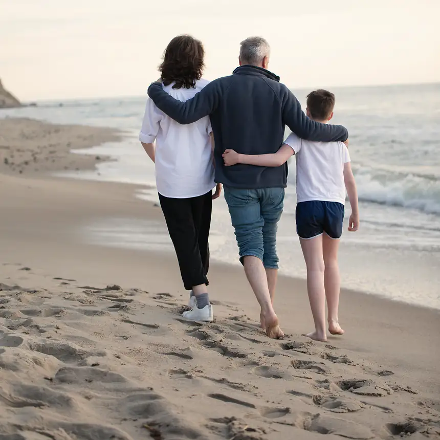A family of three walks along a quiet beach, arms around each other, seen from behind.