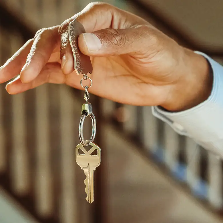 A hand holds out a single house key on a ring, a wooden staircase soft behind it.