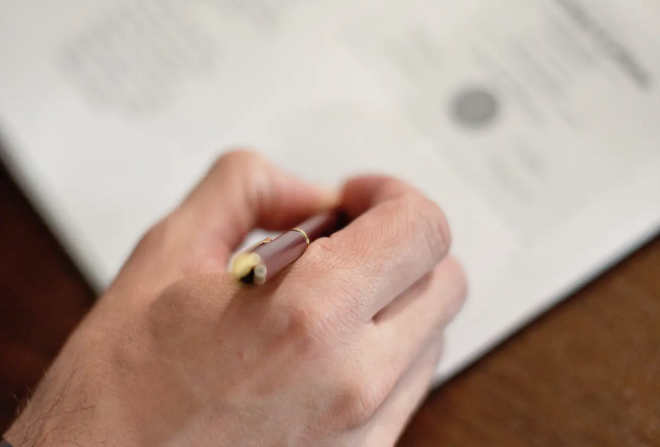 A hand signs a document with a fountain pen at a wooden table.