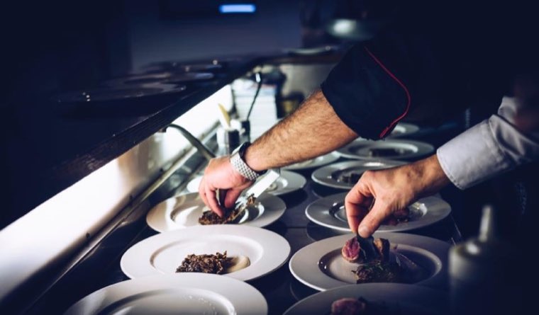 A chef’s hands placing garnish on a row of plates at the pass, under warm kitchen light.