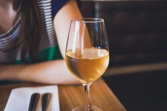 A glass of golden white wine on a wooden table next to a folded napkin and cutlery.