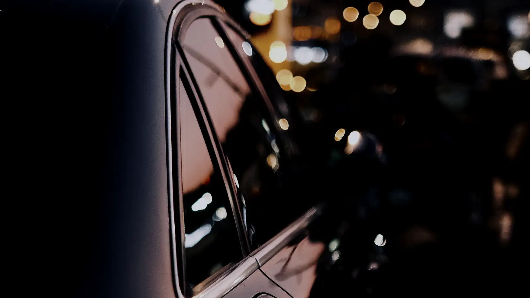 Dark tinted side windows on a black sedan reflecting the city at dusk