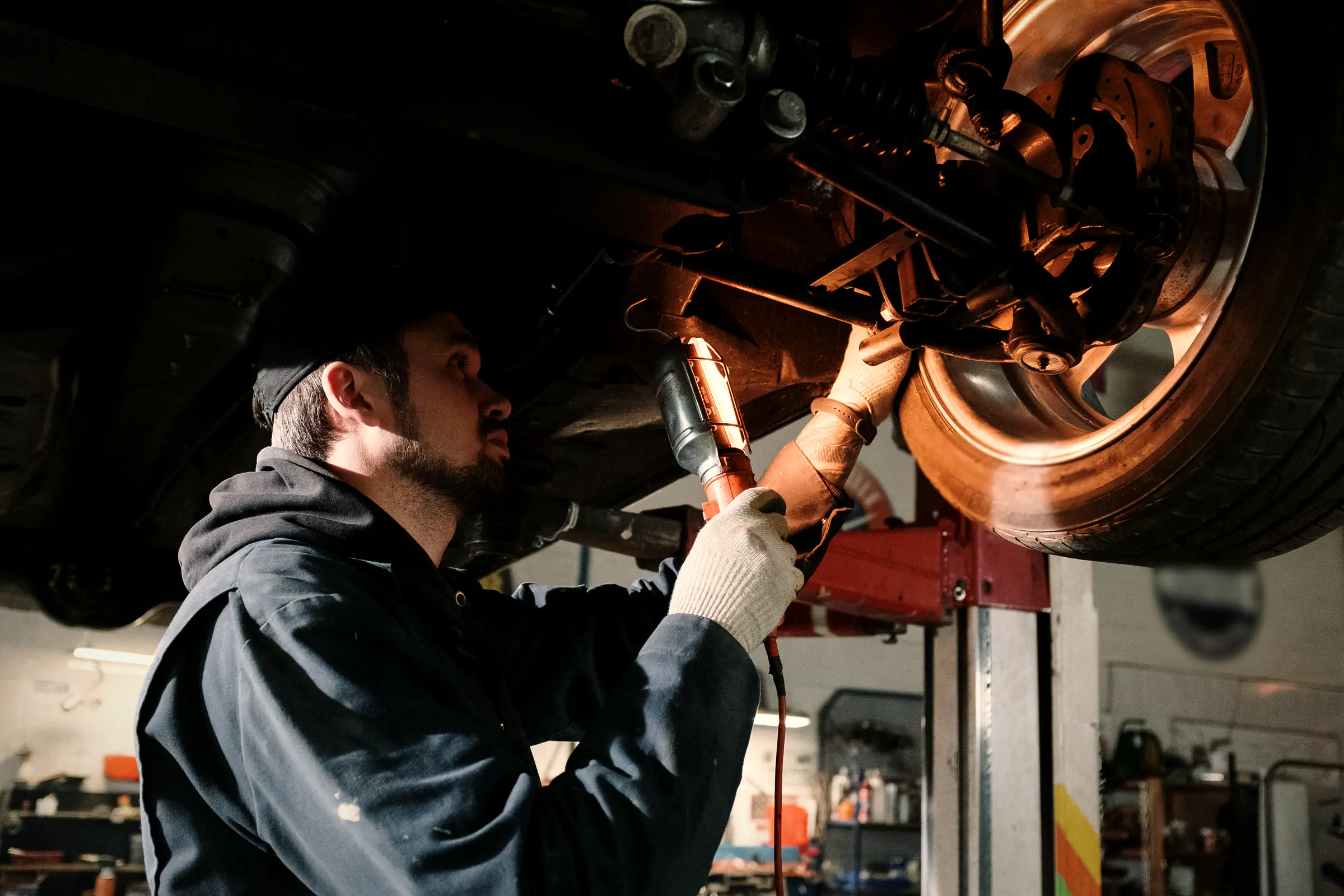 A mechanic in work gloves reaches up to the underside of a car on a lift, lit by the shop lights.
