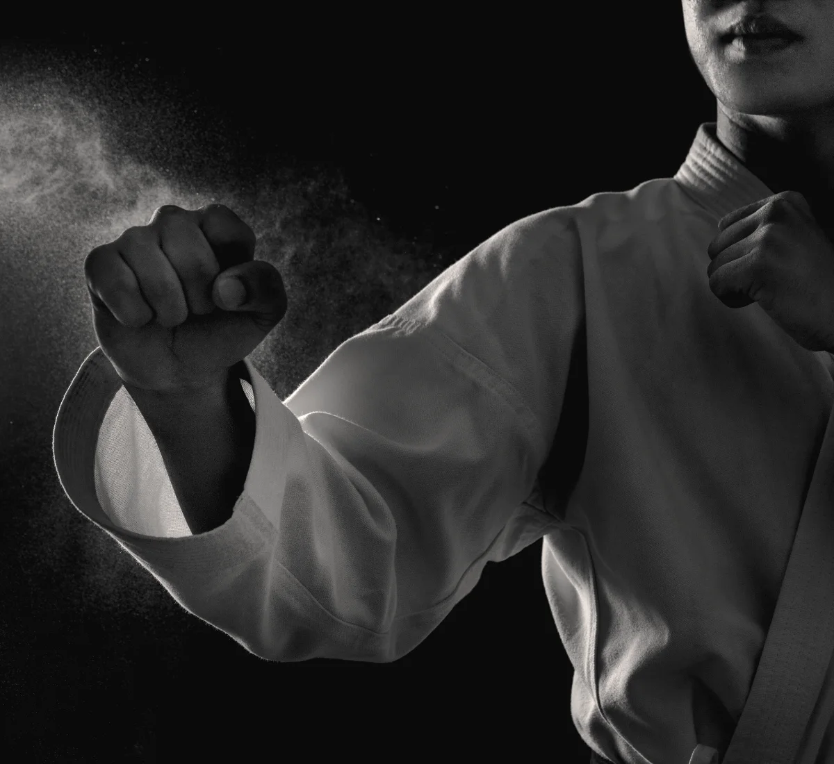 Close-up of a karateka in a white gi with both fists raised in guard, chalk dust hanging in the light