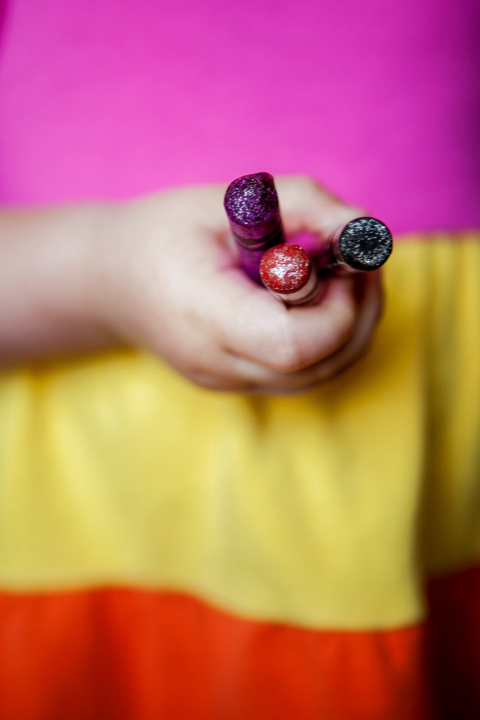 A toddler's hand holds three chunky crayons against a bright striped shirt.