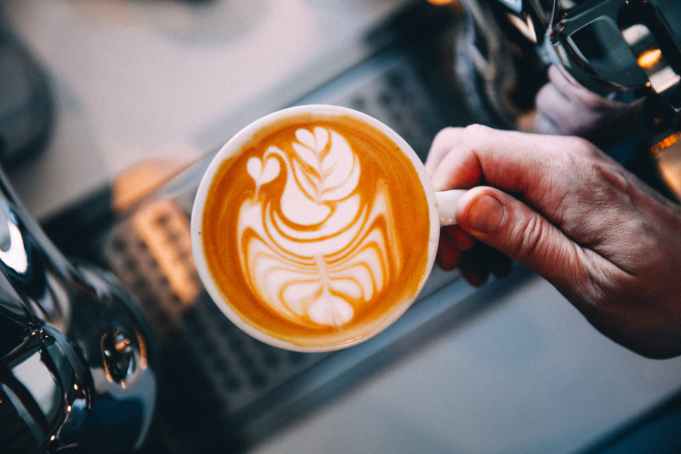 A barista holds a latte with a rosetta in the foam next to the espresso machine's steam wand.