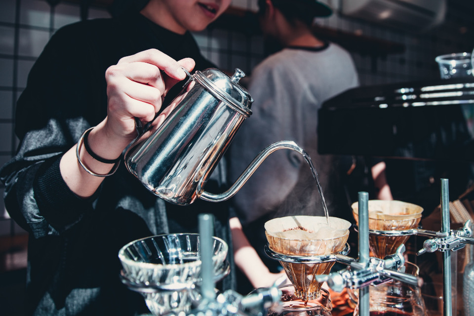 A barista pours water from a gooseneck kettle into a row of pour-over drippers.