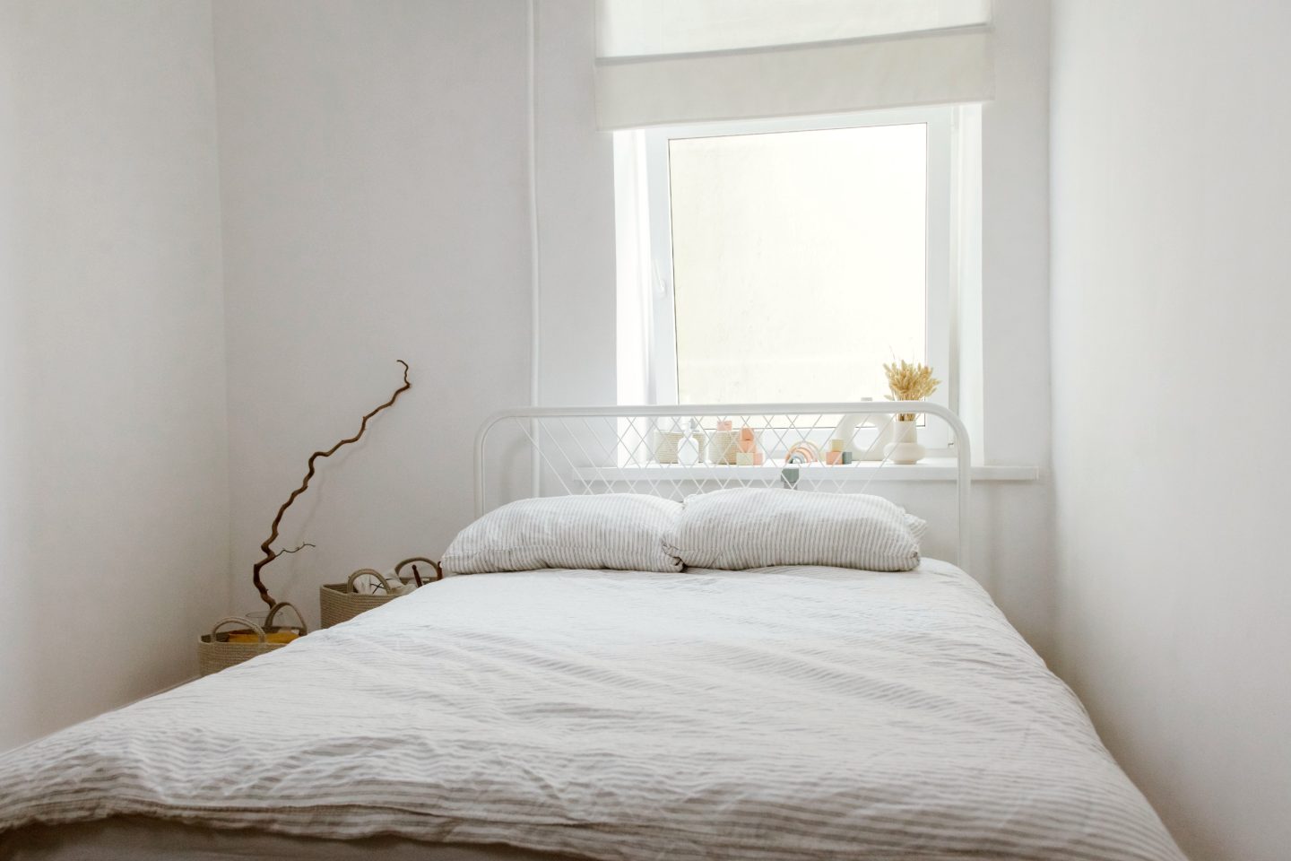 Bright bedroom with a white metal bed, grey-striped linen bedding, a twisted branch in the corner and dried grass in a vase on the windowsill