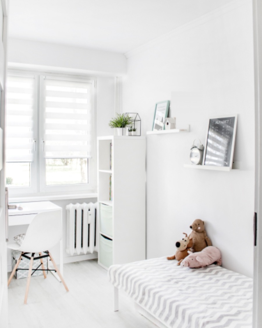 Bright white kids' room with roller blinds at the window, a white desk and chair, tidy shelves and a made bed with a few soft toys