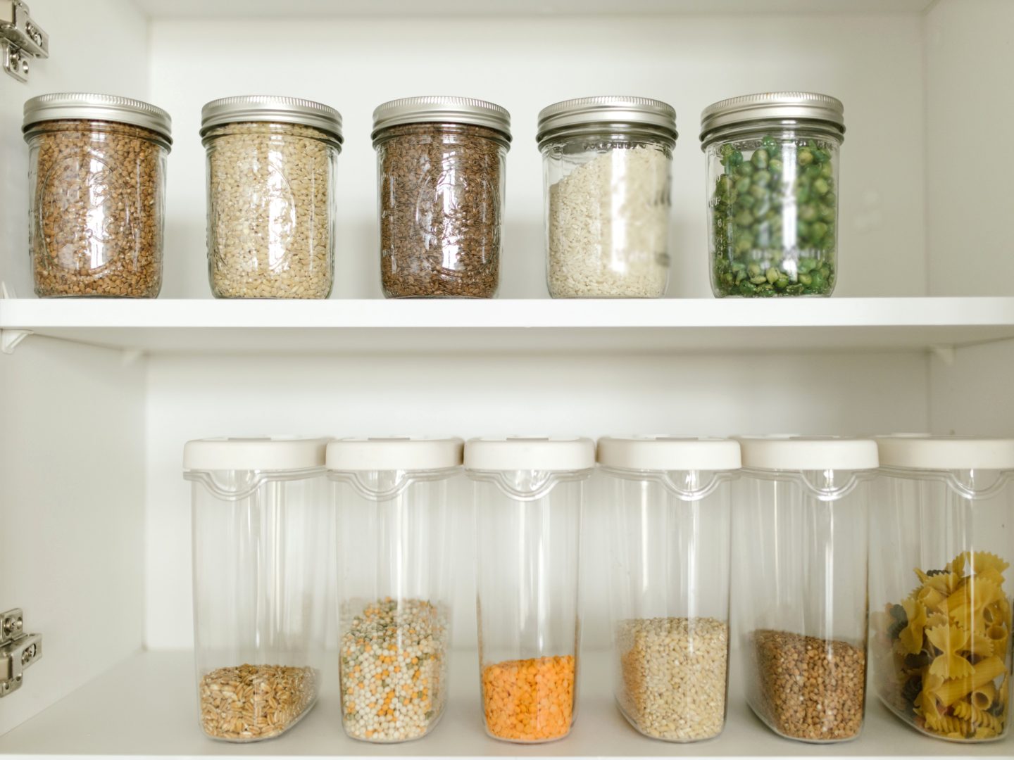 White pantry cupboard with grains and peas in clear glass jars on the top shelf, and lentils, pulses and pasta in clear containers below