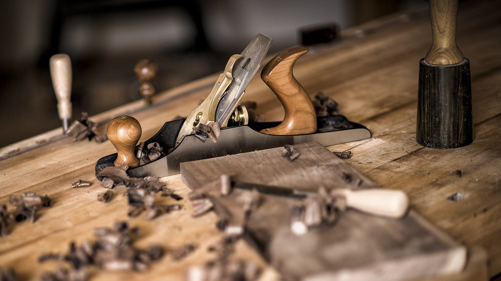 A workbench covered in shavings, with a hand plane, a chisel resting on a small board and a wooden mallet