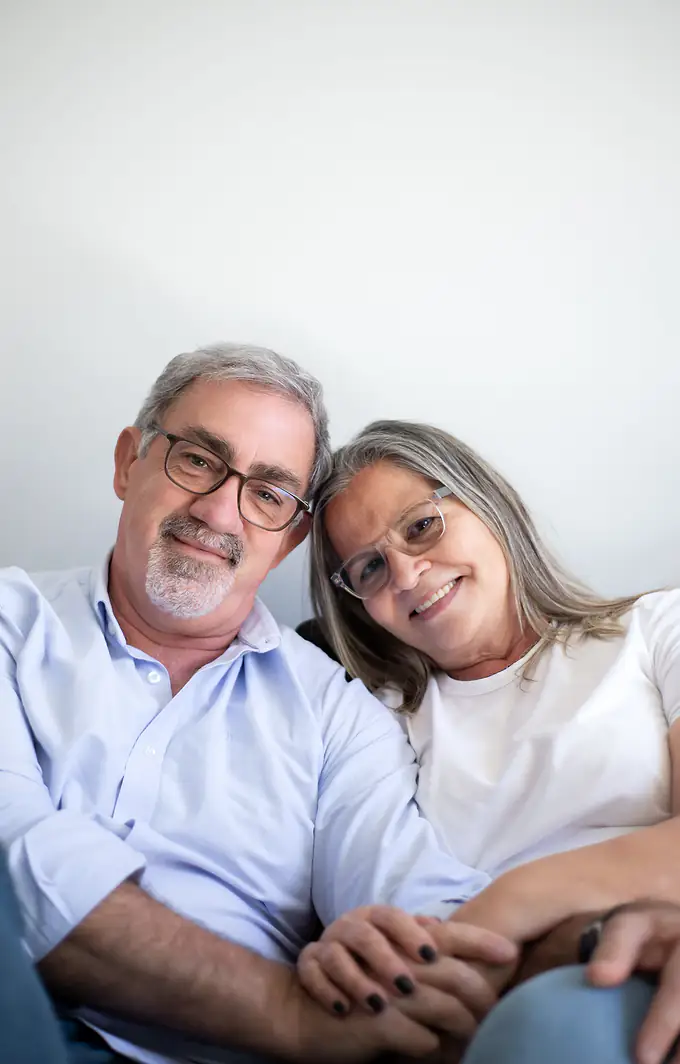 Dana and Paul, a couple in their sixties, smile together on the sofa at home.