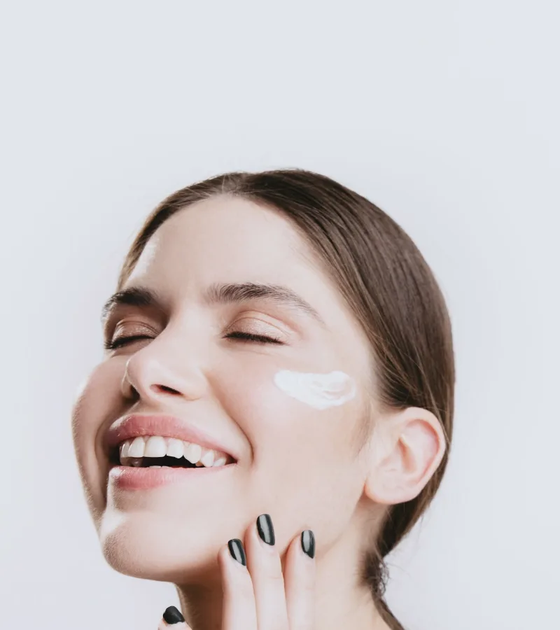 A woman laughing with her eyes closed, a dab of cream on her cheek, against a white wall