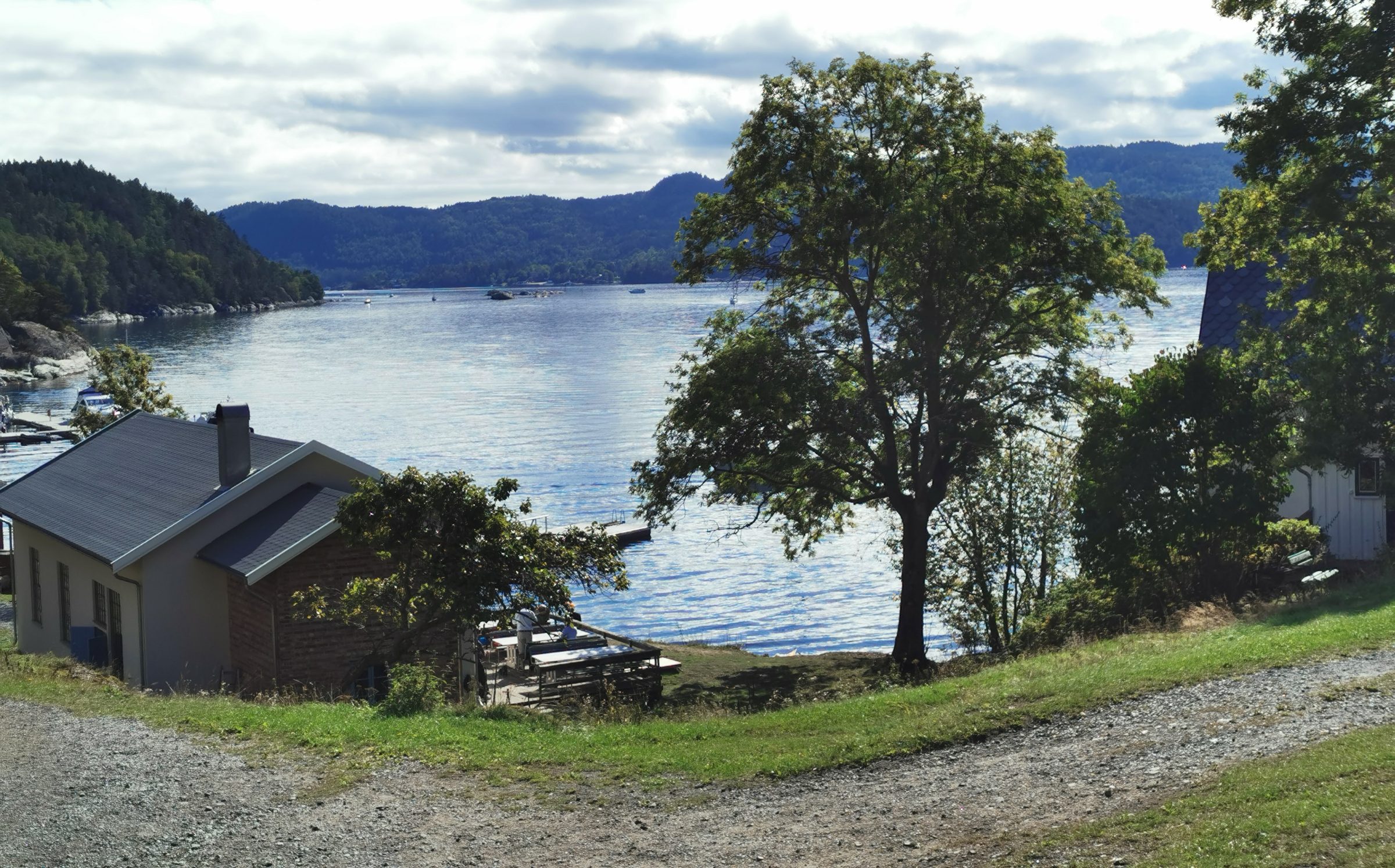 A gray-roofed house on a grassy shoreline above calm blue water, with a large tree and forested hills beyond