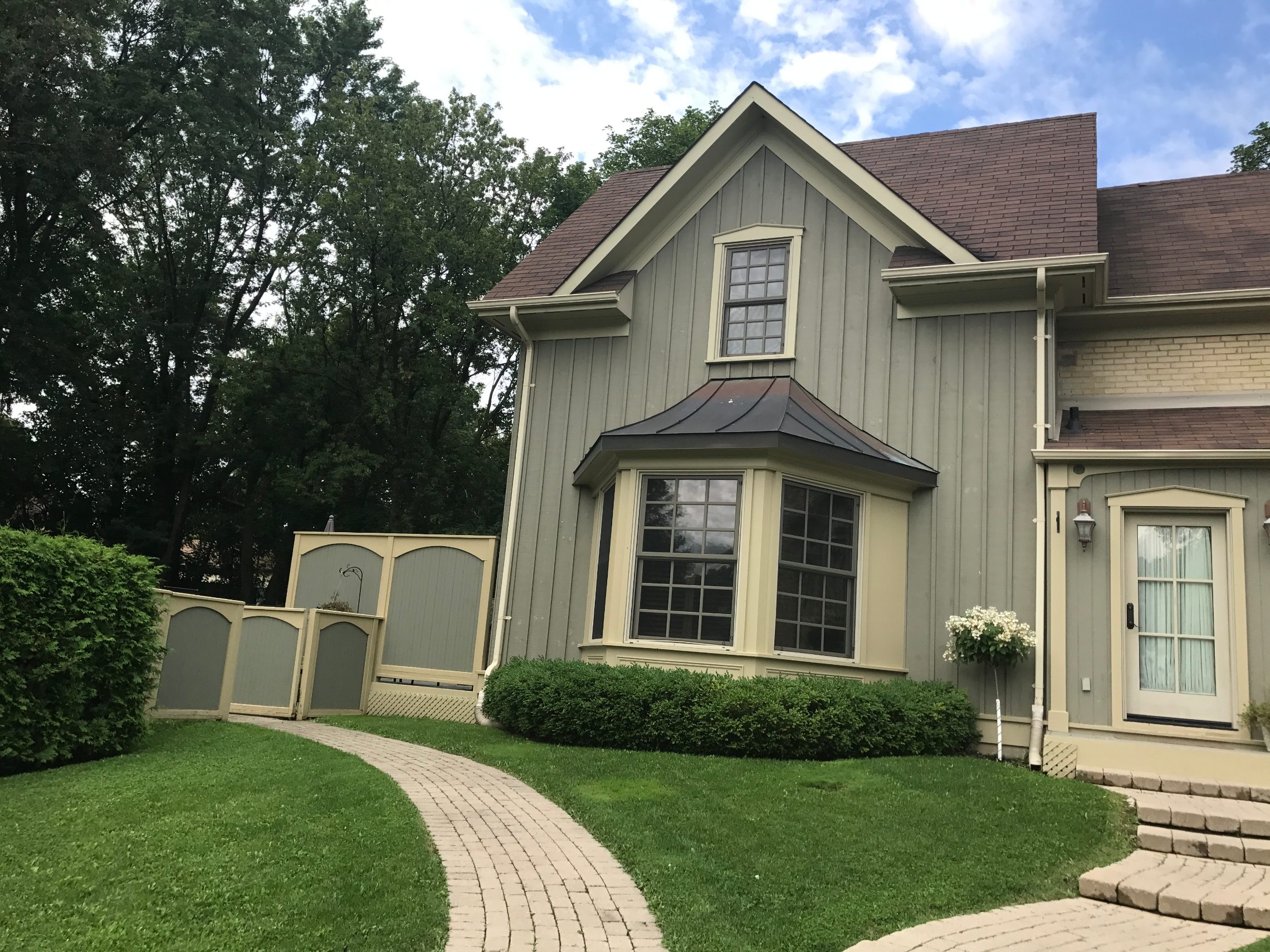Sage-green board-and-batten house with a copper-roofed bay window, clipped hedges and a curving brick path