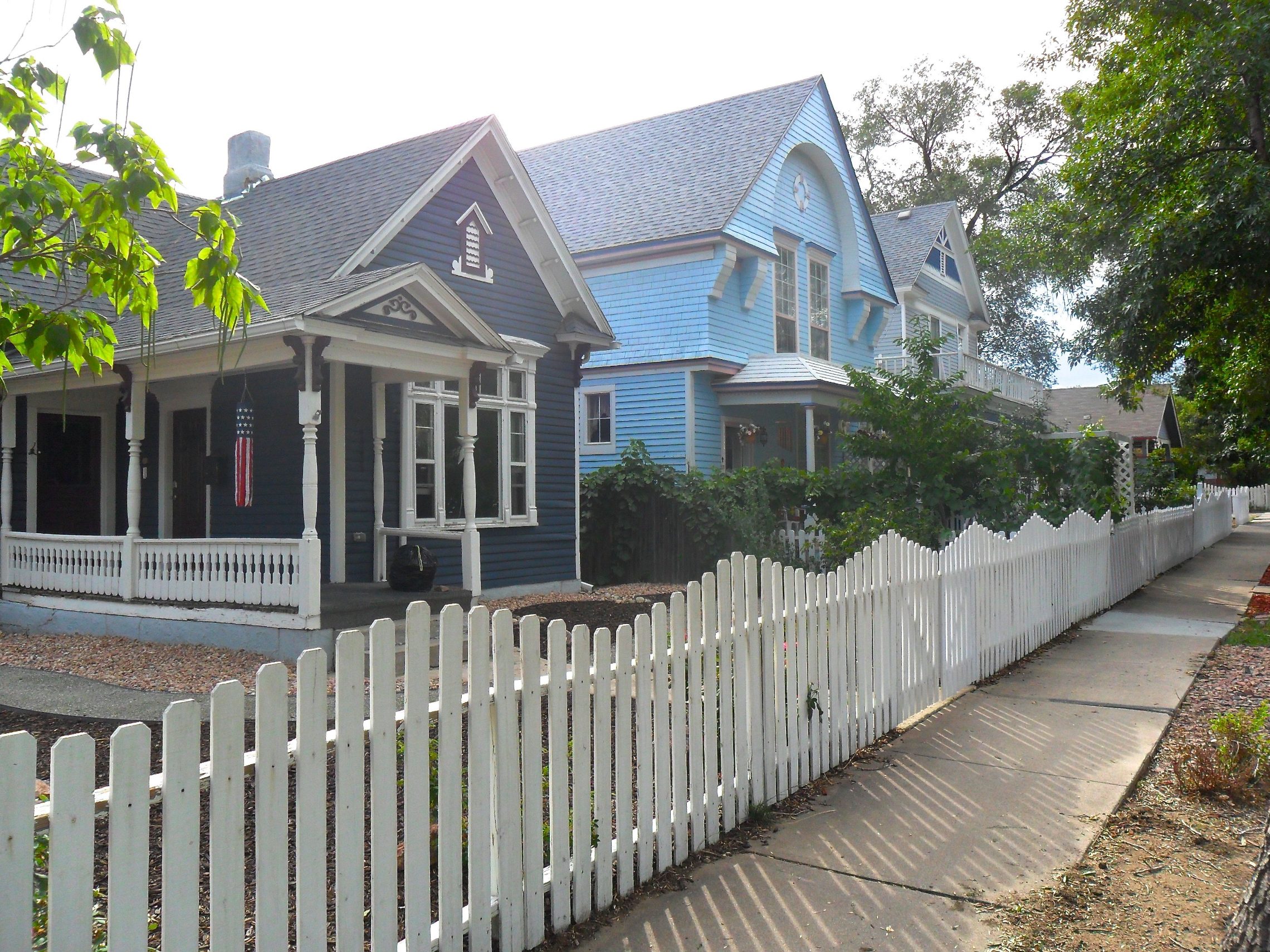 Two Victorian houses, one navy and one sky blue, with deep porches behind a white picket fence along a sidewalk