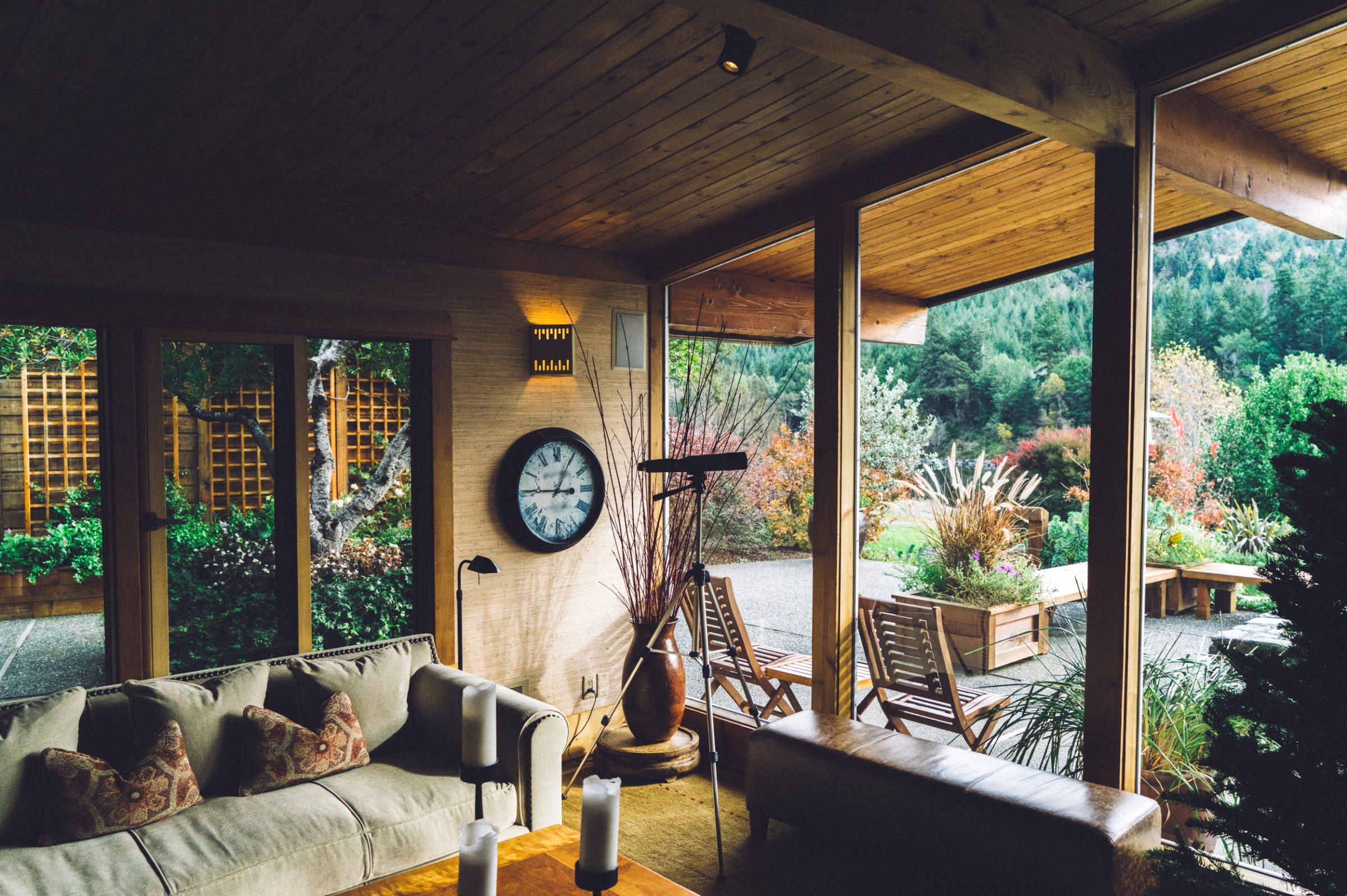 Sunroom with a wood-plank ceiling, a cream sofa and a wall clock, open on two sides to a planted garden