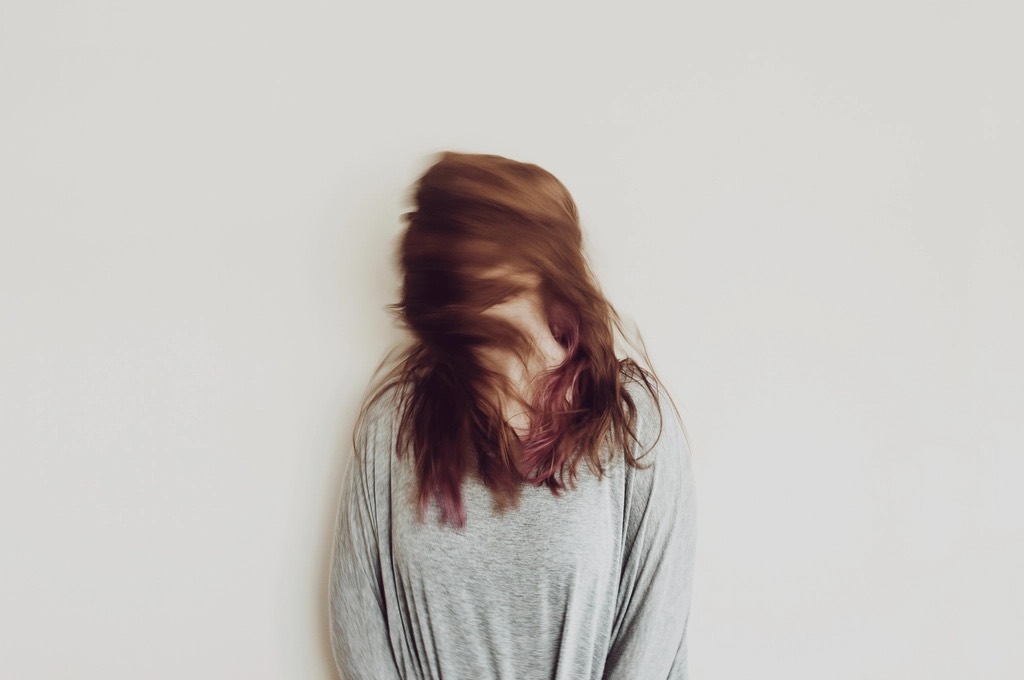 A woman with shoulder-length red-brown hair and violet ends, mid hair flip against a pale wall