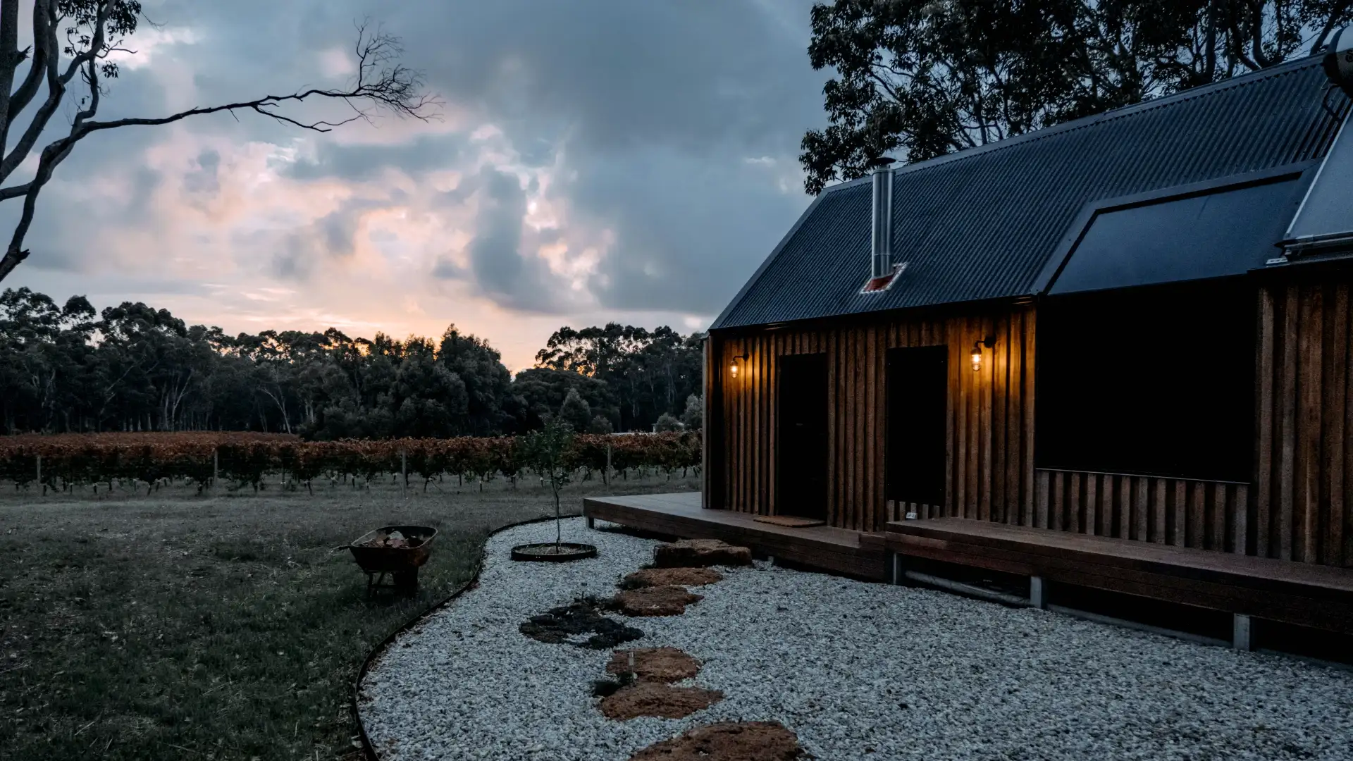 Board-and-batten timber cabin with lit wall lamps beside a gravel garden at dusk