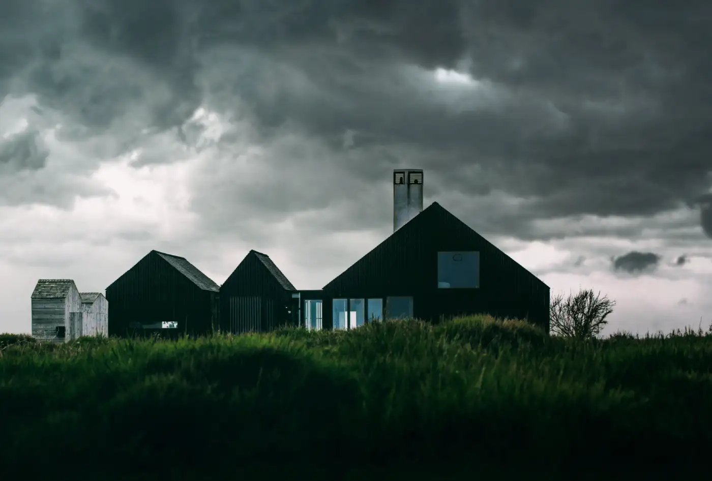 Three black-clad gabled buildings on a grassy rise under a storm sky
