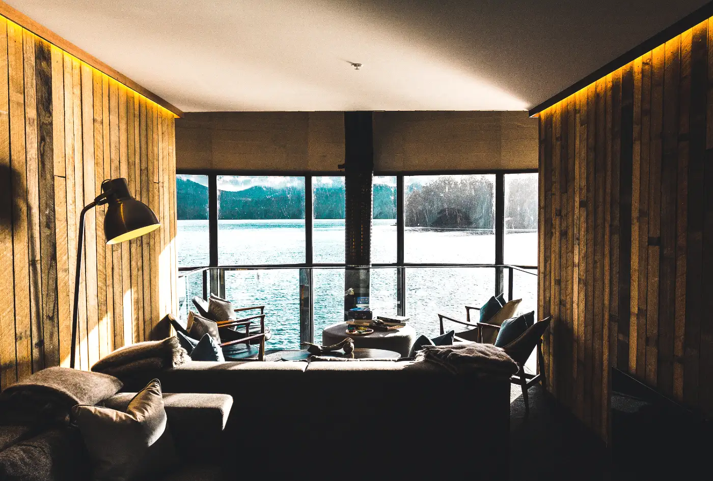 Living room with dark board walls, a lit cove ceiling and full-height windows onto a lake