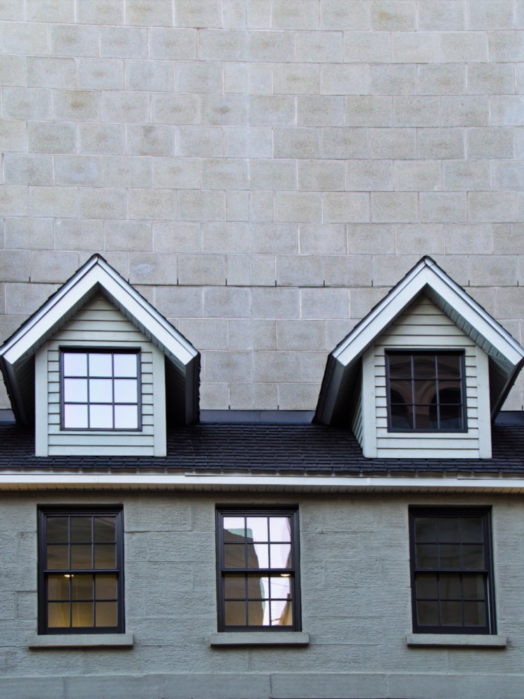 Two white dormers on a dark charcoal shingle roof above gray painted brick.
