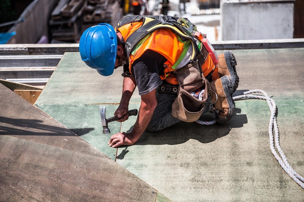 A roofer in a hard hat, hi-vis vest and harness nails down roof sheathing.