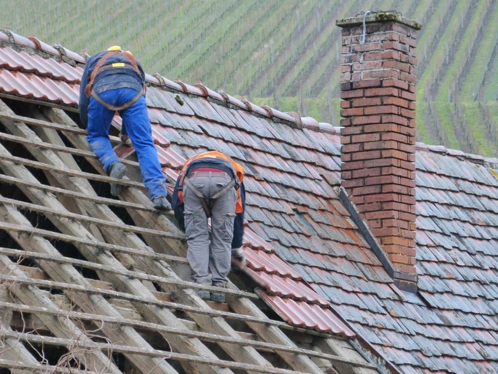 Two roofers in harnesses strip old tiles down to the battens beside a brick chimney.